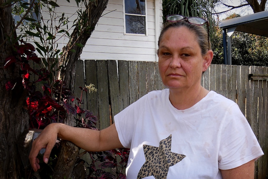 A woman in a white shirt in a backyard standing next to a tree.