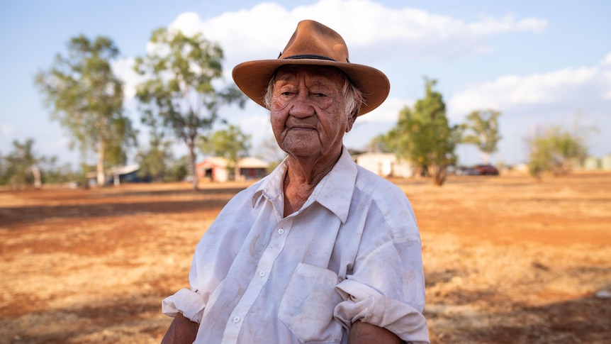 An old Aboriginal man in the outback, wearing a brown hat and white shirt with rolled-up sleeves
