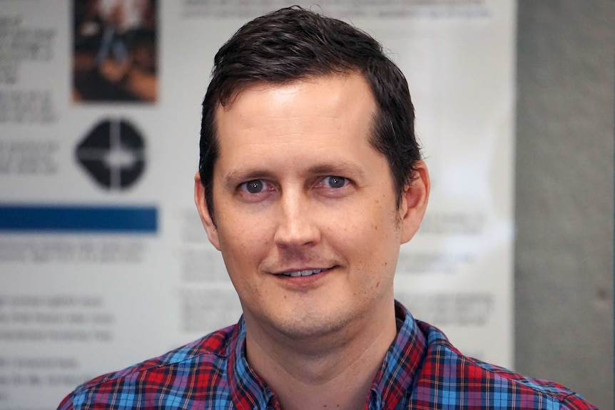 A man smiles at the camera, wearing a collared flannel shirt.