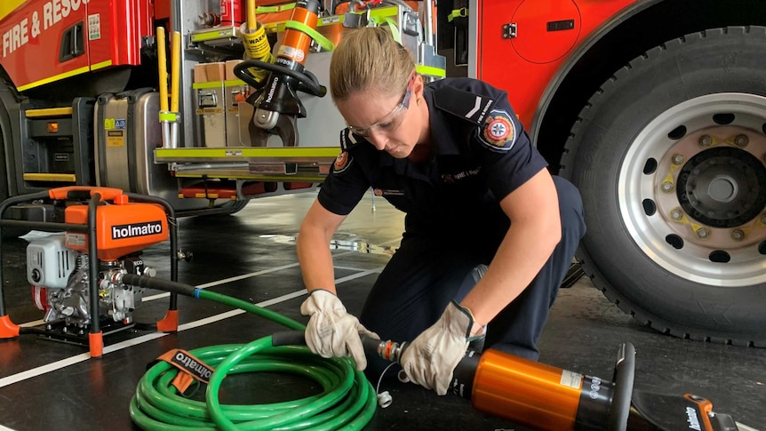 Woman in firefighter uniform kneels on ground and looks at equipment.