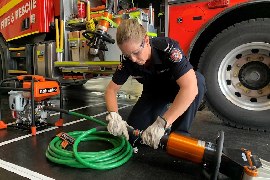 Woman in firefighter uniform kneels on ground and looks at equipment.