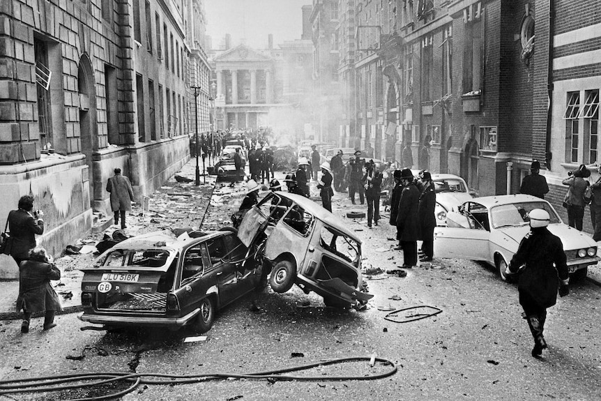 A black and white photo shows two bombed-out cars on top of each other with debris strewn across the road.