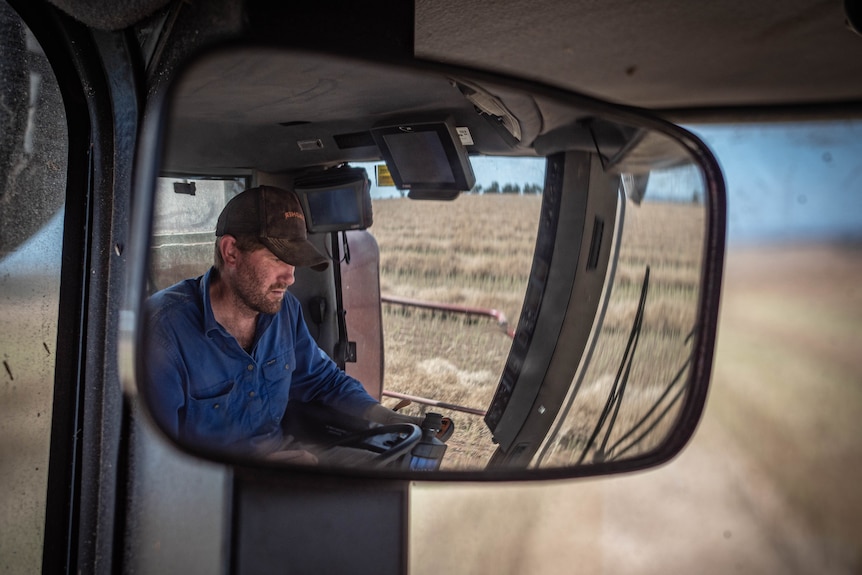 A man wearing a blue shirt, brown cap, seen through a rear view mirror in the cabin of a header.