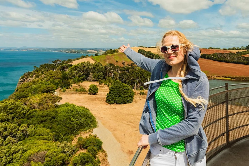 A woman gestures to the horizon while standing on the deck of a lighthouse.