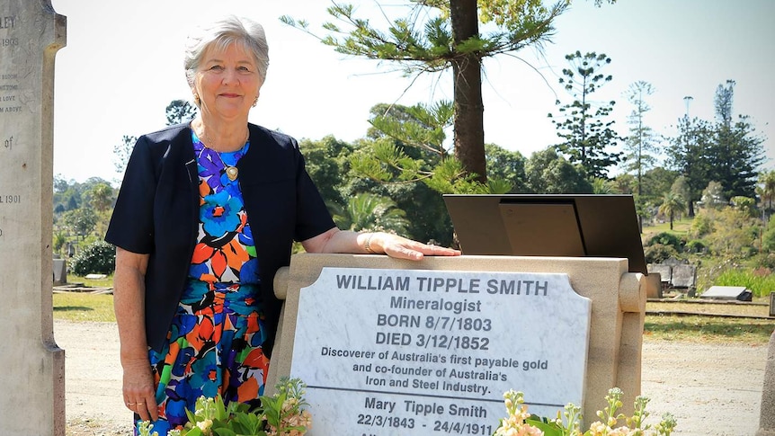 An elderly woman at a gravesite with her hand on a headstone.