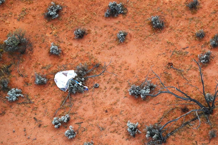 An aerial view of the space capsule and its parachute surrounded by red dirt and bushes