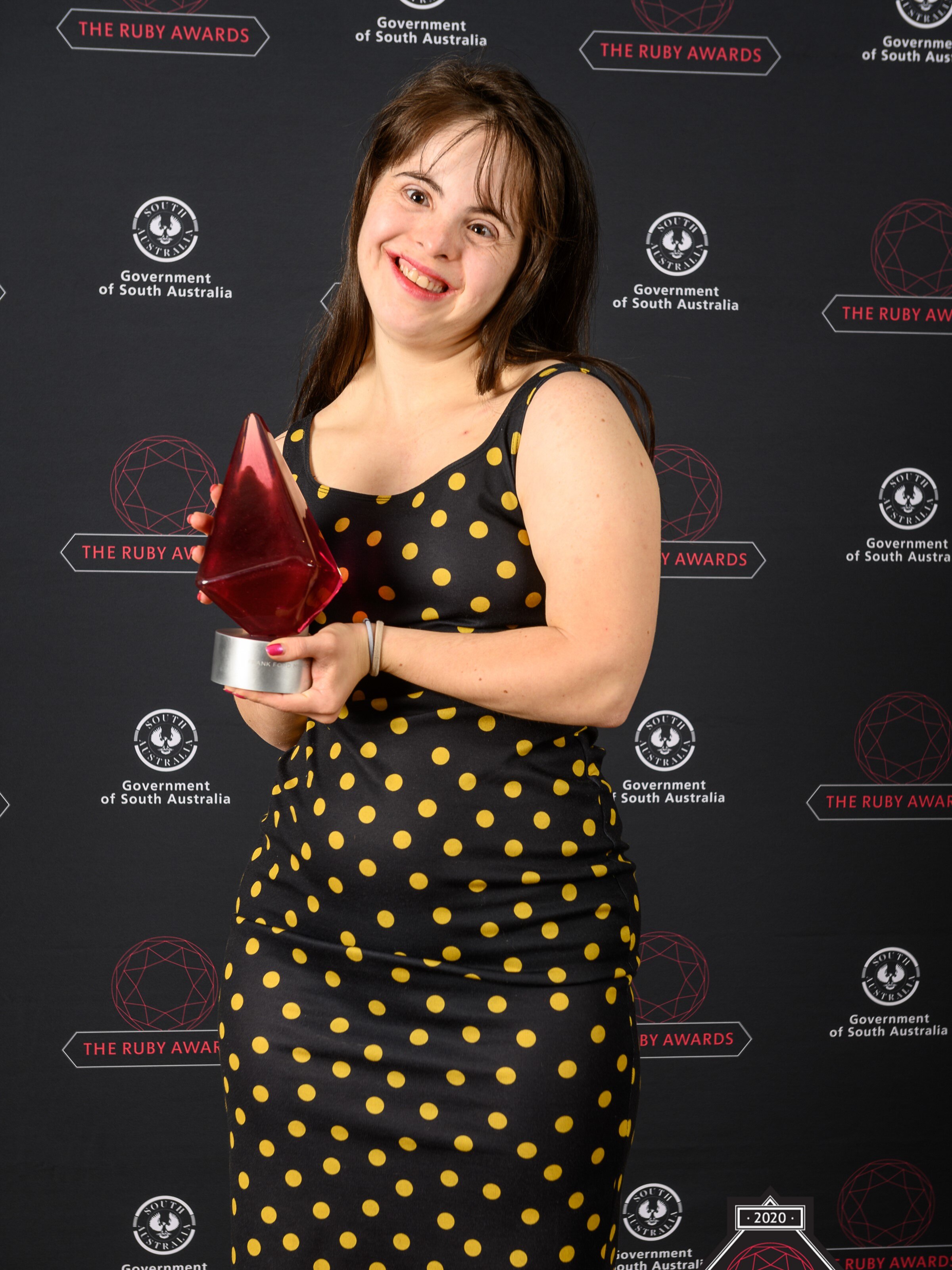 Photo of a dancer posing with a trophy - with brown hair, wearing a black dress with yellow dots, holding a red glass trophy