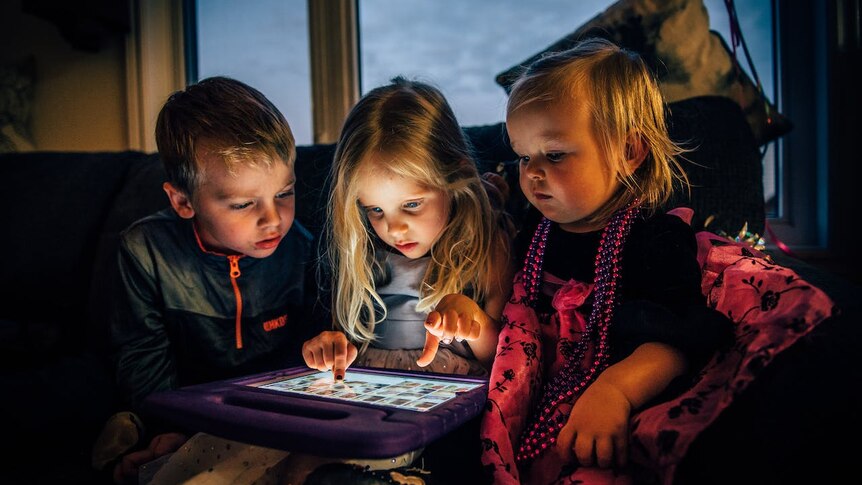Three young children stare down at a tablet computer on their lap.