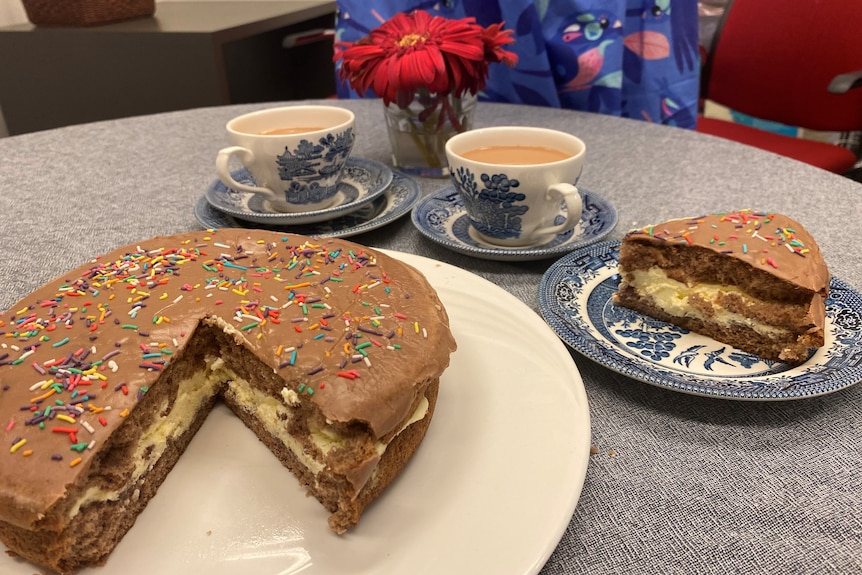 A ginger fluff sponge sits on a table with two cups of tea. One slice has been cut and sits on a small plate.