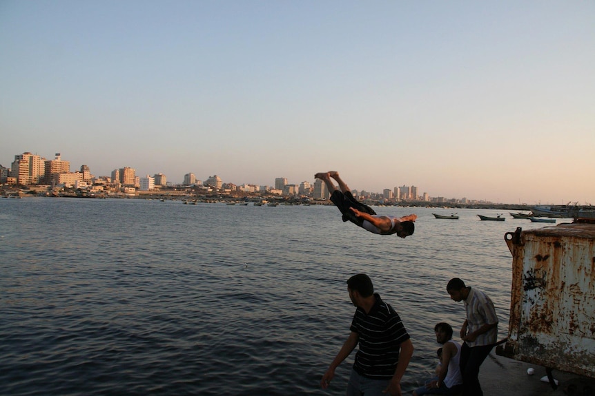 A wide photo of a man mid-air while doing a flip into the sea off a port at dusk, as other men stand nearby.
