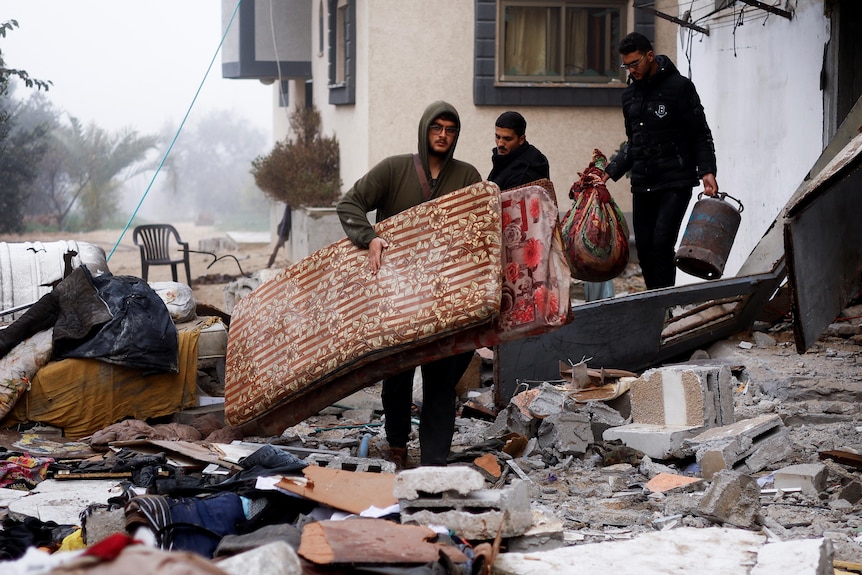 Men carrying mattresses are surrounded by debris from an air strike. 