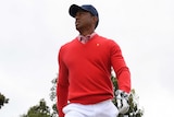 Tiger Woods walks on the course, carrying a club. A large crowd of people stands behind him.