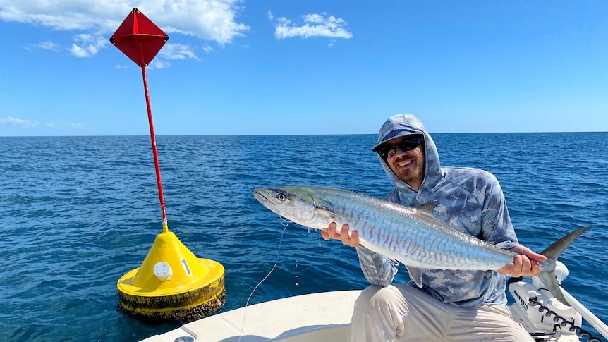 A man holding a caught Spanish Mackerel on a boat in front of a yellow buoy in the water.