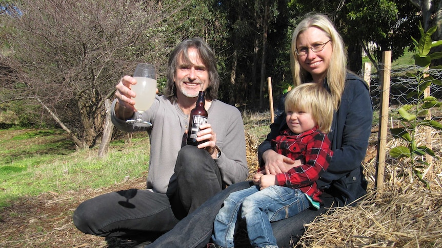 A family sit in a fruit tree orchard holding a glass of ginger beer