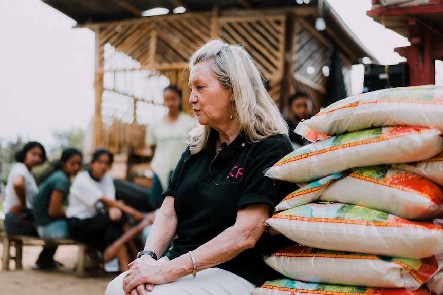 A women with blond hair and a black top sits next to large bags of rice. A group of Balinese people are behind her