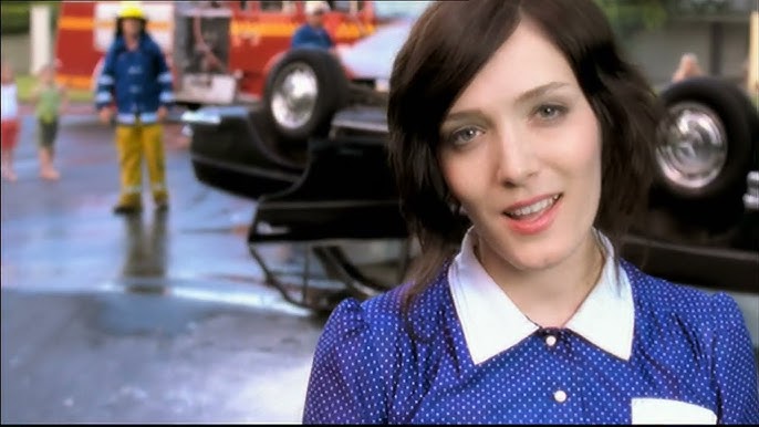 Una persona con una camiseta azul de lunares se encuentra frente a un coche volcado y un equipo de rescate.