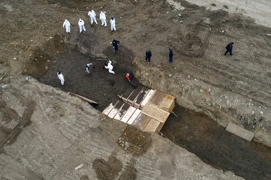 workers in white and black protective clothing dig a large grave with coffins inside