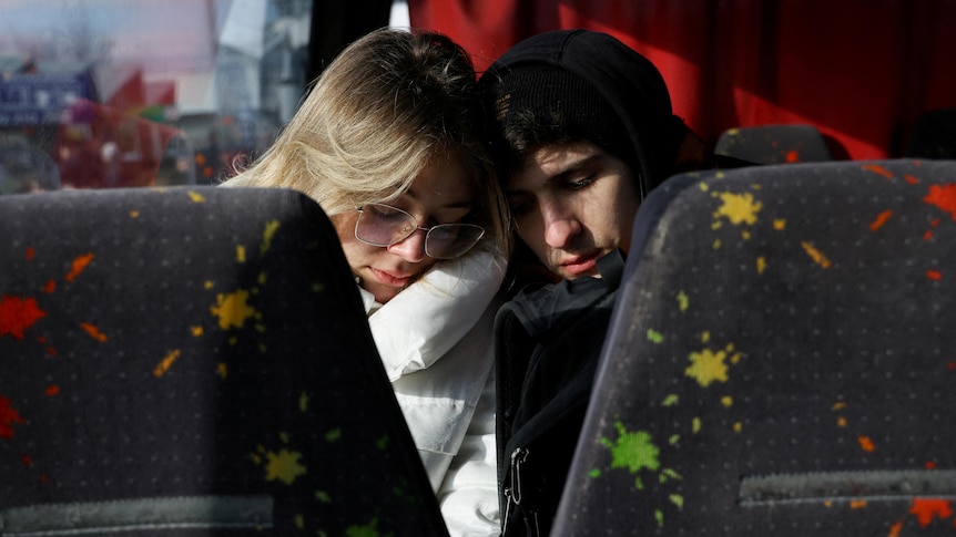 a man and a woman sit on a bus with their eyes closed leaning in to each other with bus seats in the foreground