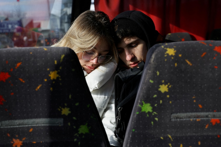 a man and a woman sit on a bus with their eyes closed leaning in to each other with bus seats in the foreground