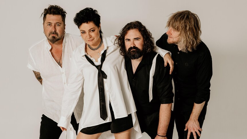 Four members of The Superjesus stand arm-in-arm, side-by-side in a white photo studio. Two wear white tops, two wear black.