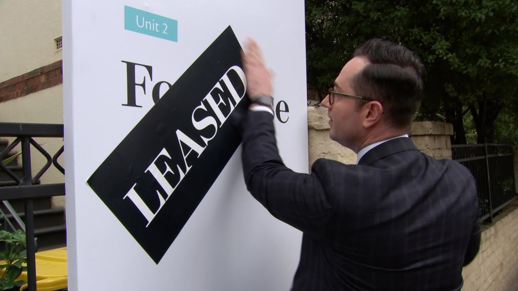 A man in a suit places a large sticker saying 'Leased' on a real estate sign.