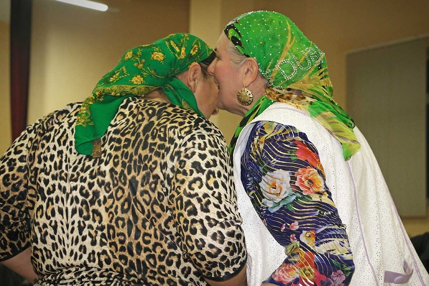 Two older gypsy women wearing head scarves whisper into each other's ears.