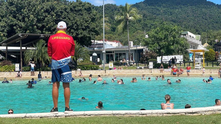 A man in a red lifeguard shirt, with his back to the camera, watching over people swimming at a lagoon.