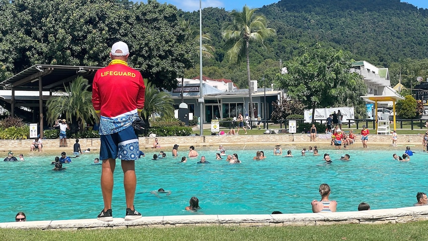 A man in a red lifeguard shirt, with his back to the camera, watching over people swimming at a lagoon.