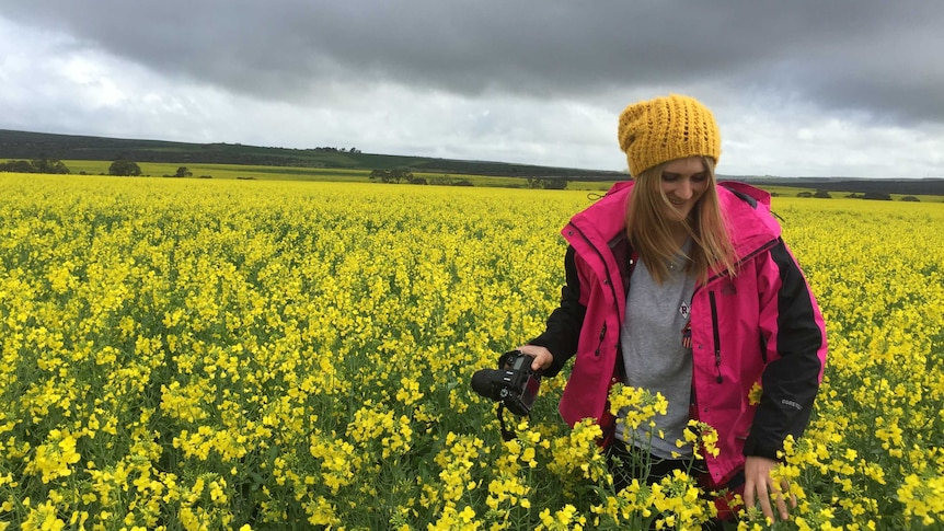 A girl standing in a yellow canola crop under a cloudy sky holding a camera