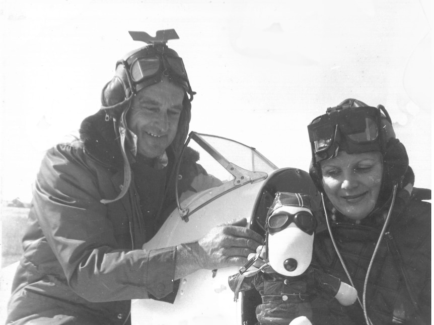 On old photo of a couple gathered by a small plane. The woman is holding a stuffed animal