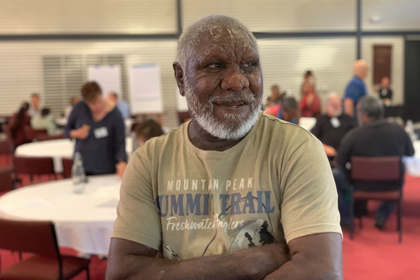 A portrait shot of an older Aboriginal man with short gray hair and a beard, he has his arms folded and is wearing an olive colo