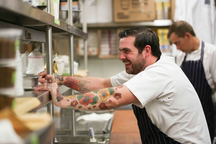 A man in an apron stands in a kitchen, resting his hands on some metal shelves.