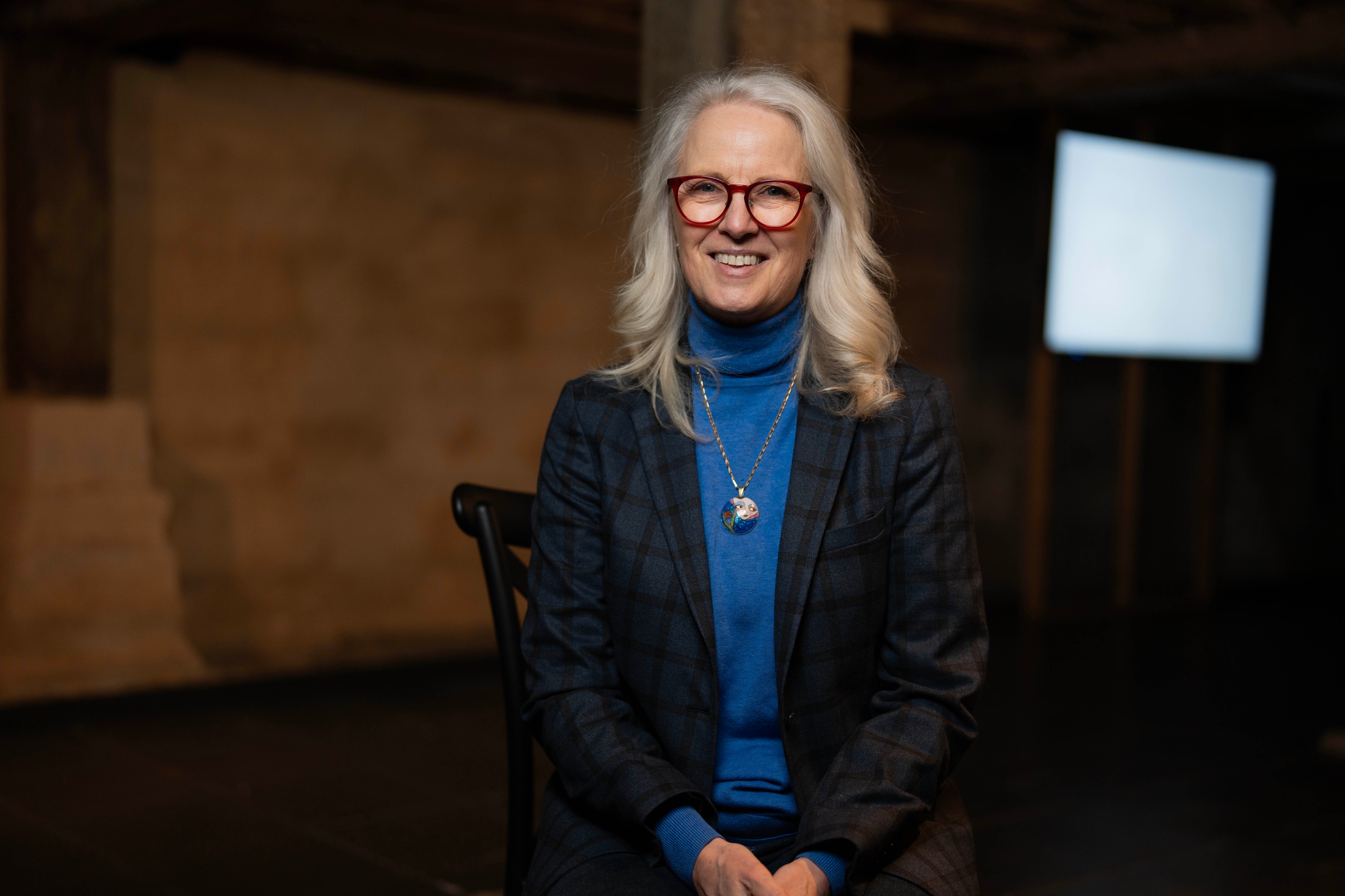 A lady in a blue top and blazer sitting in a museum looking at the camera.