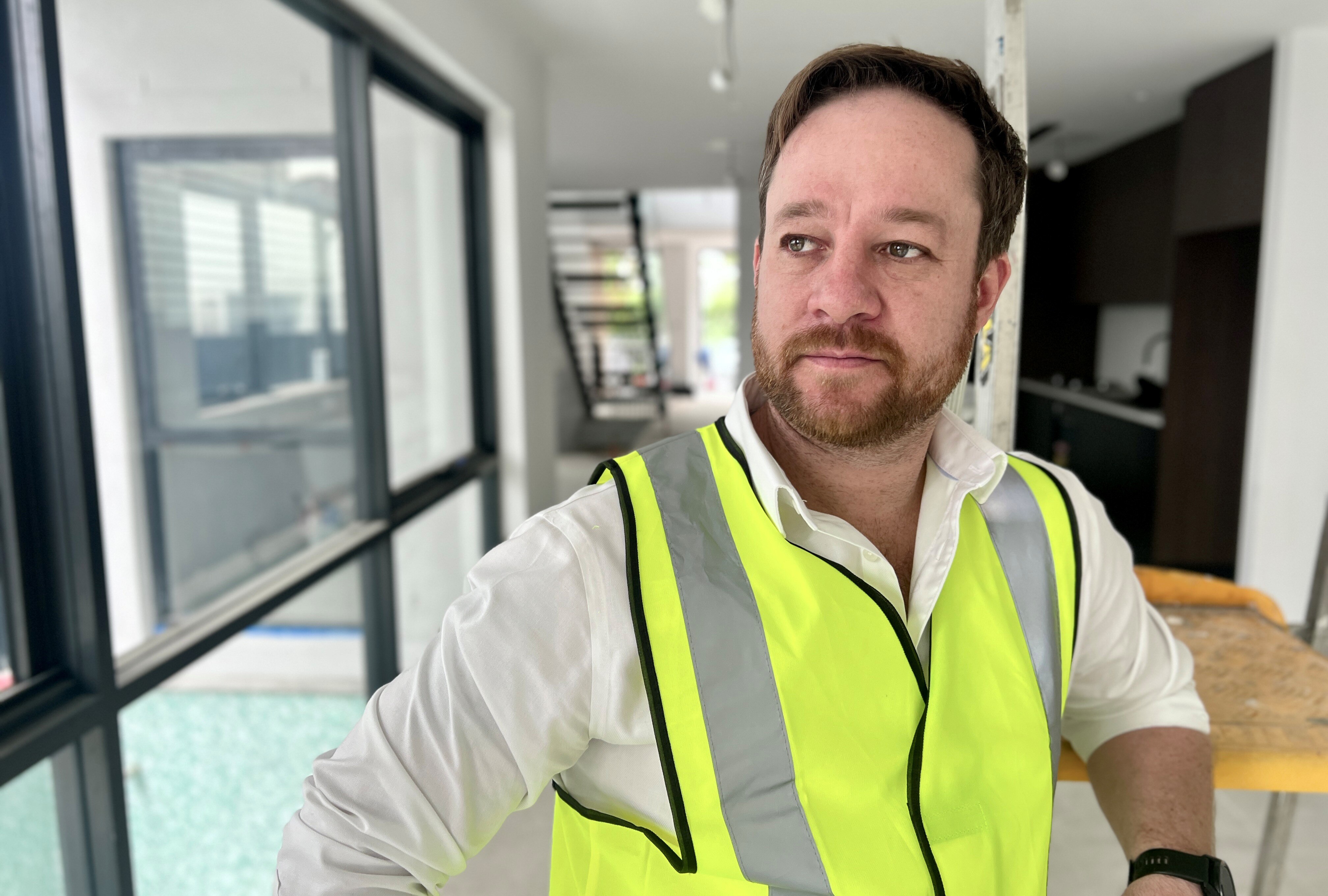 Serious Matthew has a beard and moustache, hands on hips, wears high-viz jacket over white shirt, in a house under construction.