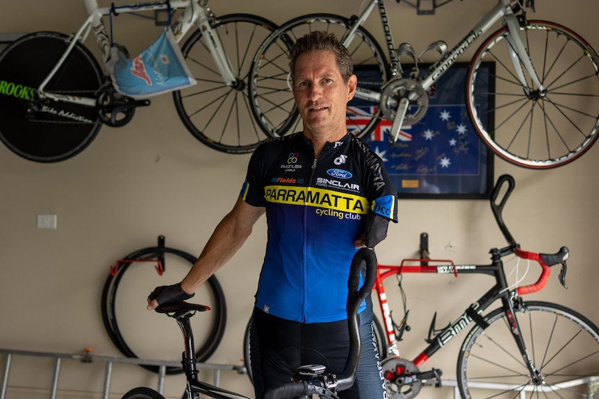 A man in cycling attire stands in a shed with bikes hanging on the wall behind him.