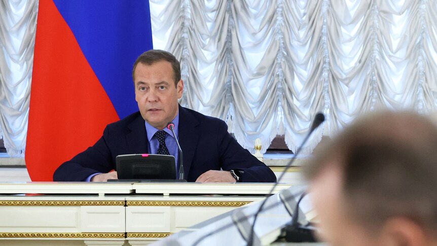 medvedev sits at a table in front of a microphone with the russian flag behind him 