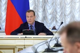 medvedev sits at a table in front of a microphone with the russian flag behind him 