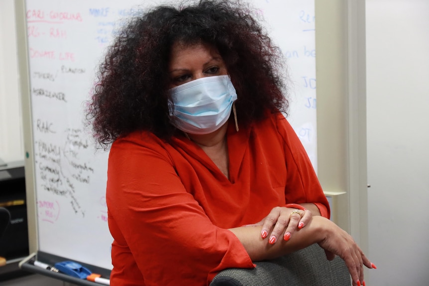 A woman in a red shirt wears a face mask and leans on a chair. 