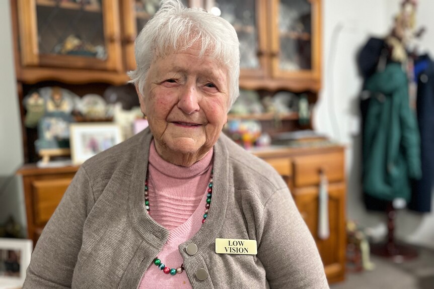 White haired lady wearing a grey cardigan, a beaded necklace and a 'low vision' tag, smiles at camera 