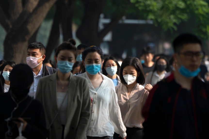 People, mostly wearing masks, walk along a path past trees during peak hour.