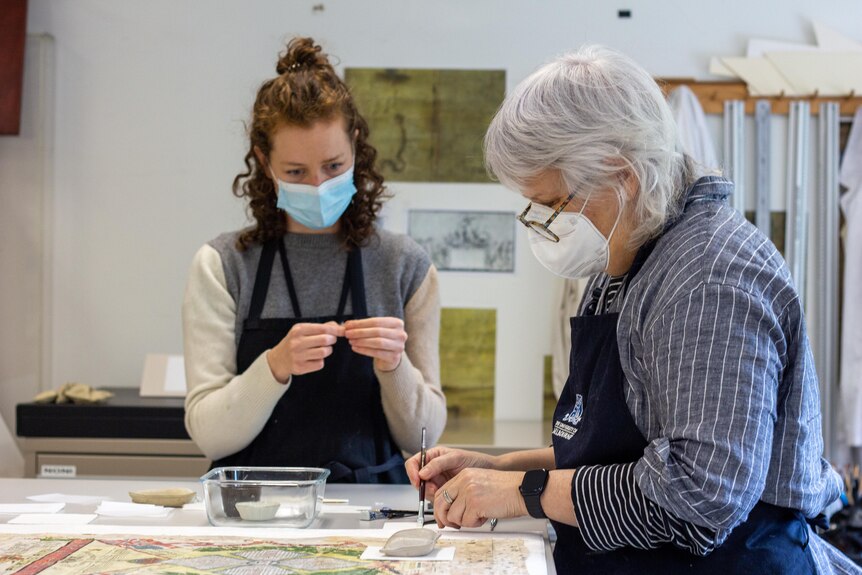 Two masked students with aprons on work to restore torn paper on the nominal roll.