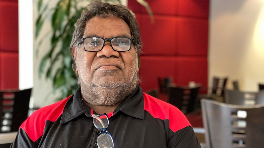 A man wearing reading glasses and a black and white shirt looks seriously at the camera in a restaurant