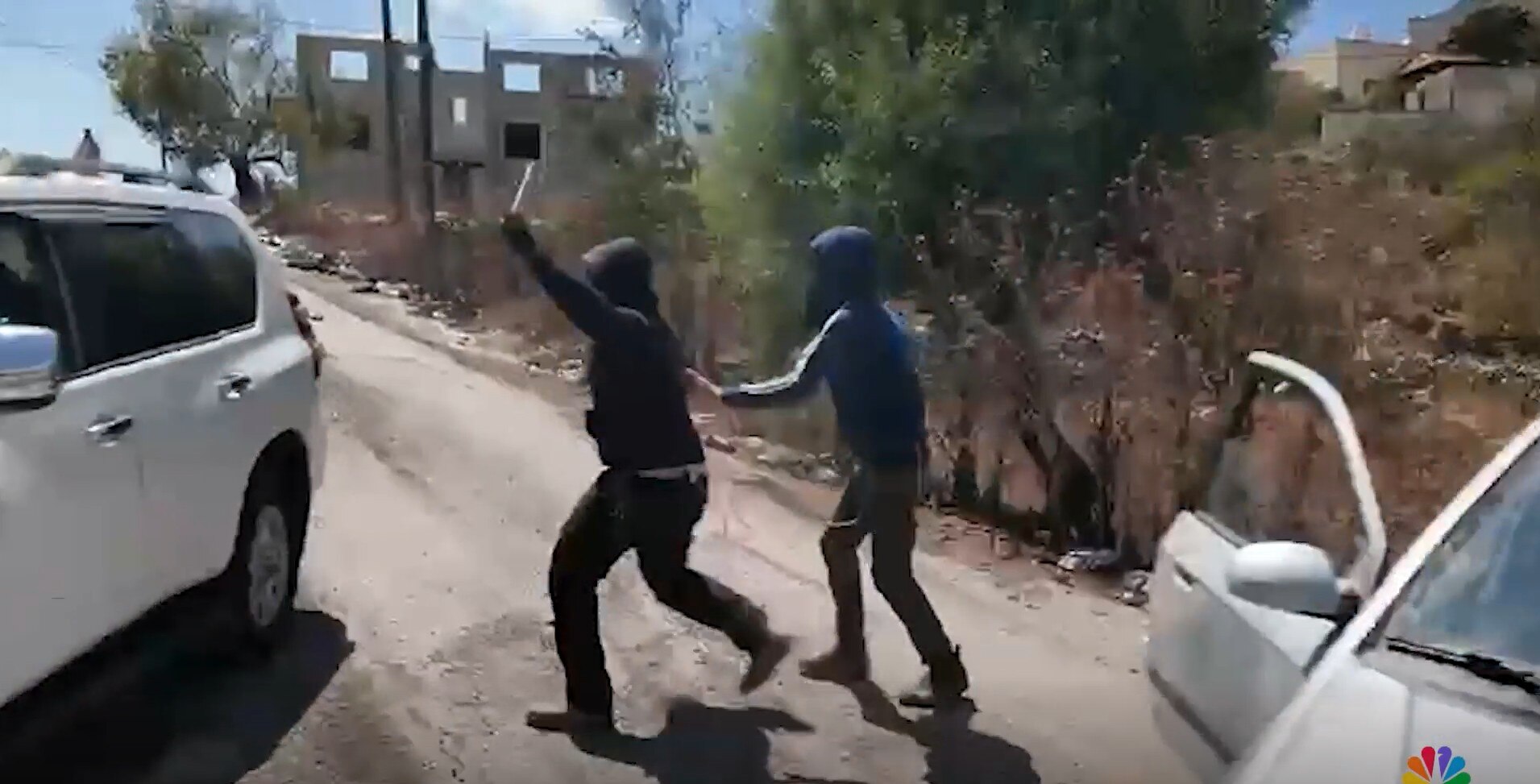 A screenshot from a video showing two men in hoods and masks approaching a car with sticks raised.