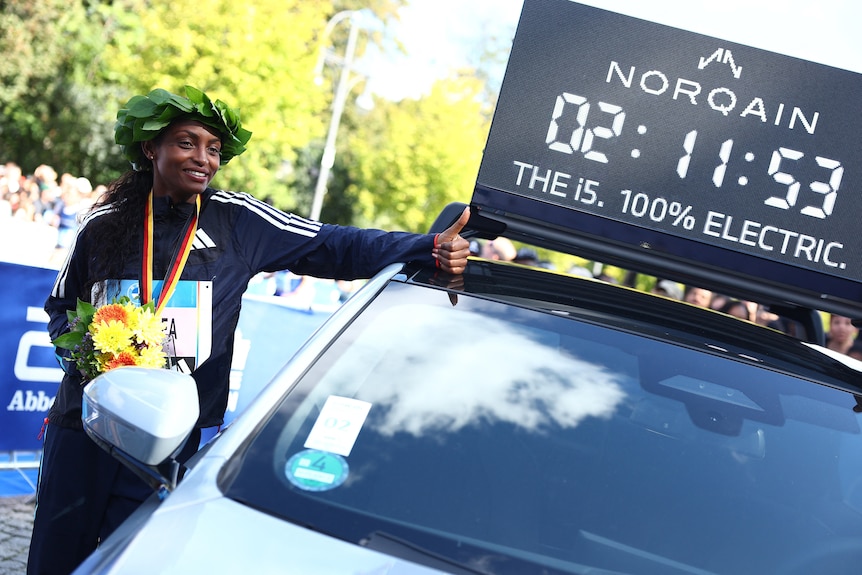 A black woman wearing a medal around her neck gives thumbs up next to timer on top of car in street