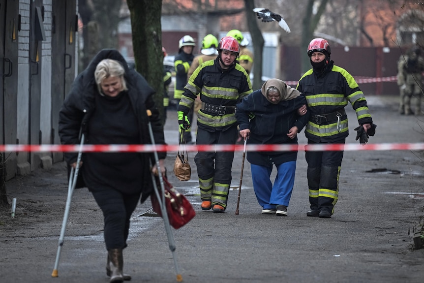 Rescuers help a older woman with a walking stick near a residential building
