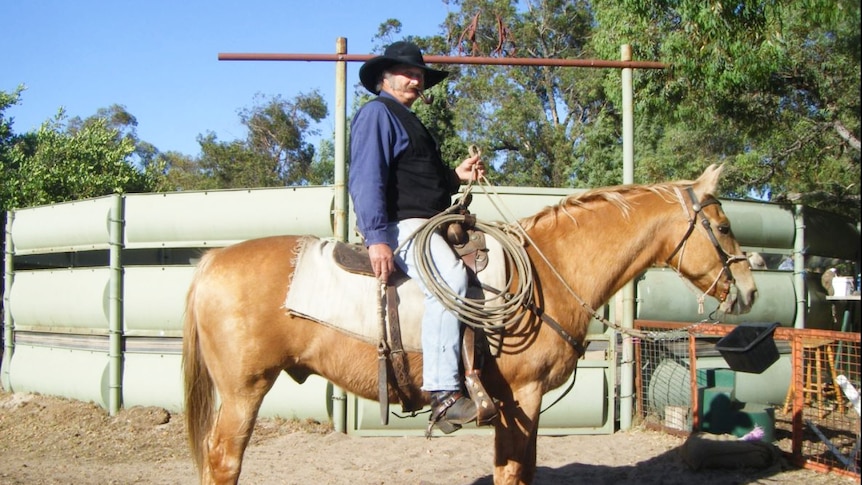 A man in a wide brim hat and smoking a tobacco pipe sits on a horse on a rural property