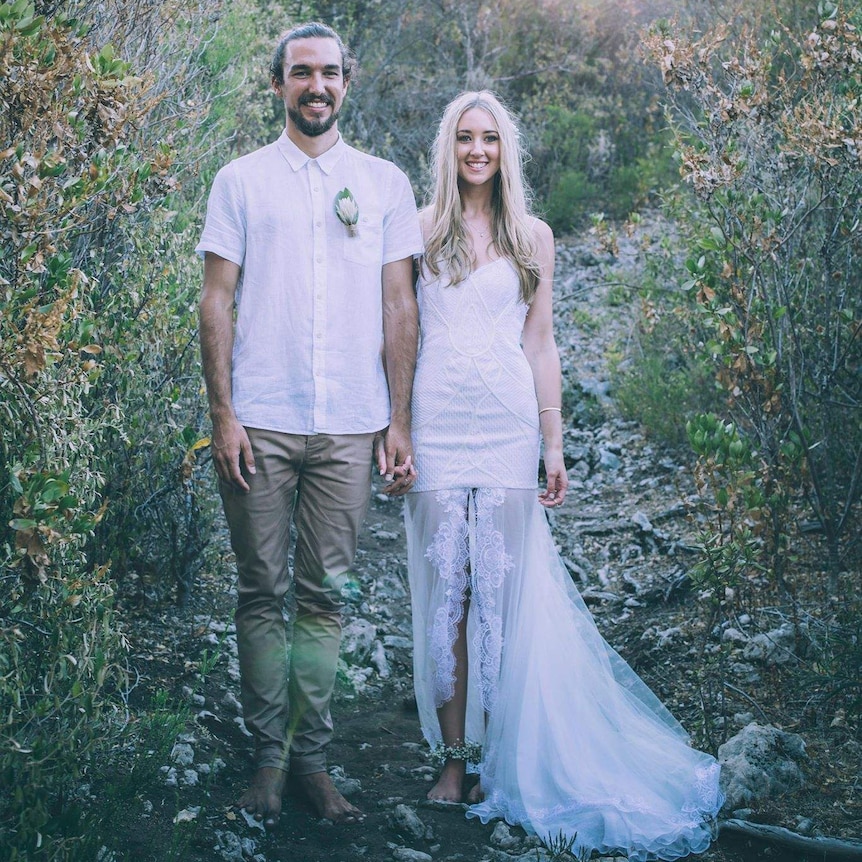 A bride and and groom standing next to each other wearing no shoes. The groom is in rolled-up chino pants.