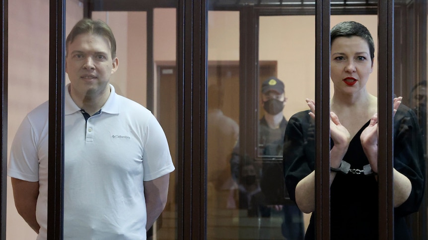 A man and a woman stand handcuffed, side-by-side, in a small jail cell in the middle of a court room