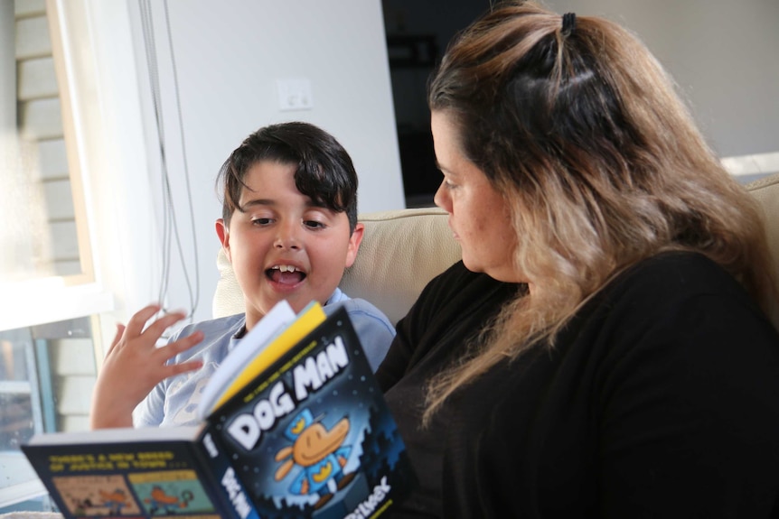 A young boy reads a children's book with his mother, sitting on a yellow couch.