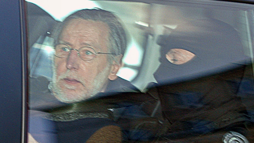 A grey-haired man sits next to a police officer wearing a balaclava